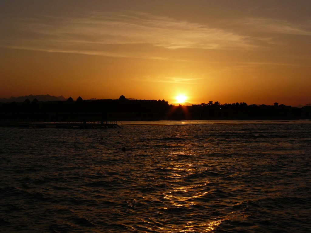 Ein Sonnenuntergang über dem Meer, mit einer Silhouette der Küste und Palmen im Vordergrund. Das Bild zeigt die Küste von Hurghada, Ägypten.